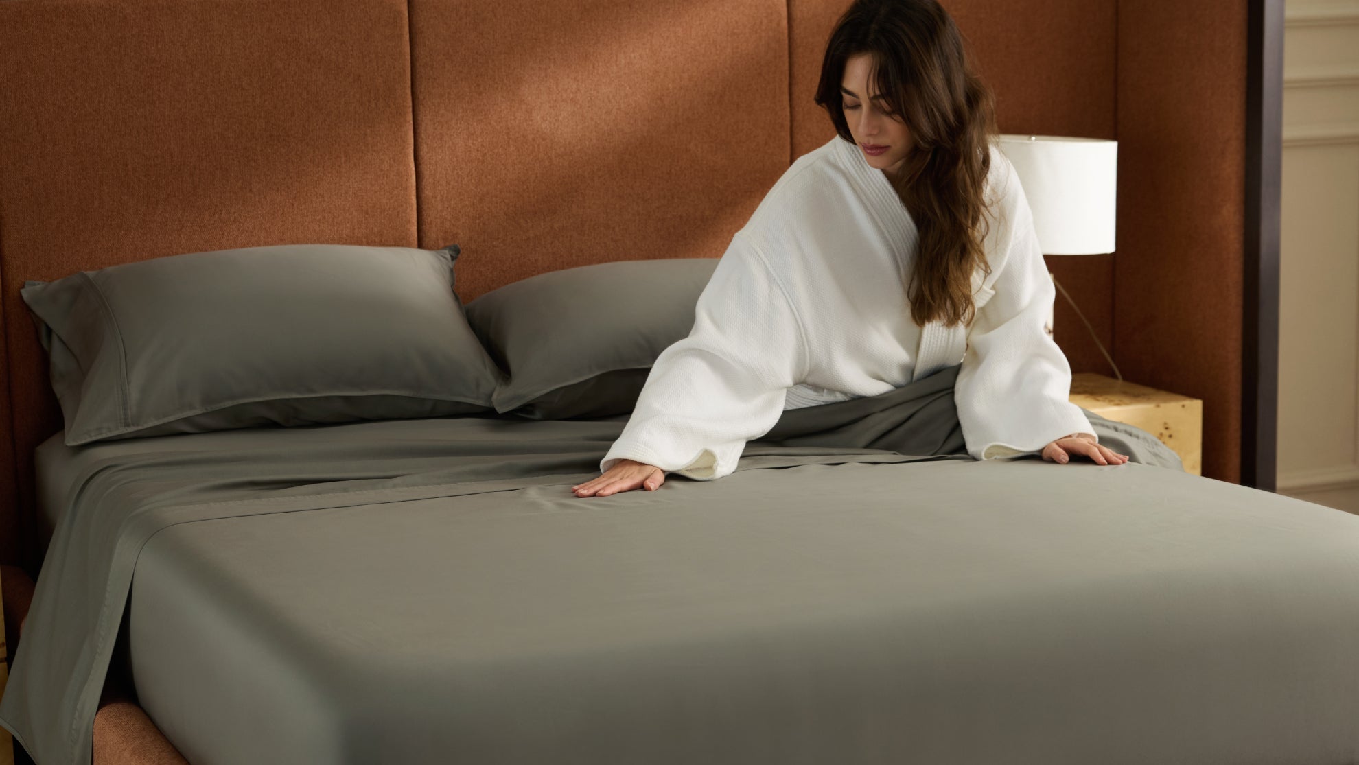 Woman smoothing out fresh grey cotton bed sheets in a bedroom, ensuring the fabric is clean and stain-free.