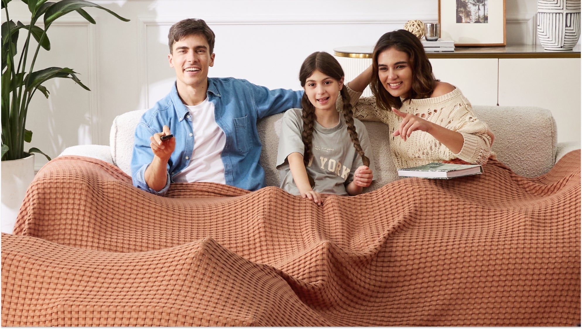 Family of three watching TV under a cozy waffle blanket during a home Winter Olympics watch party.