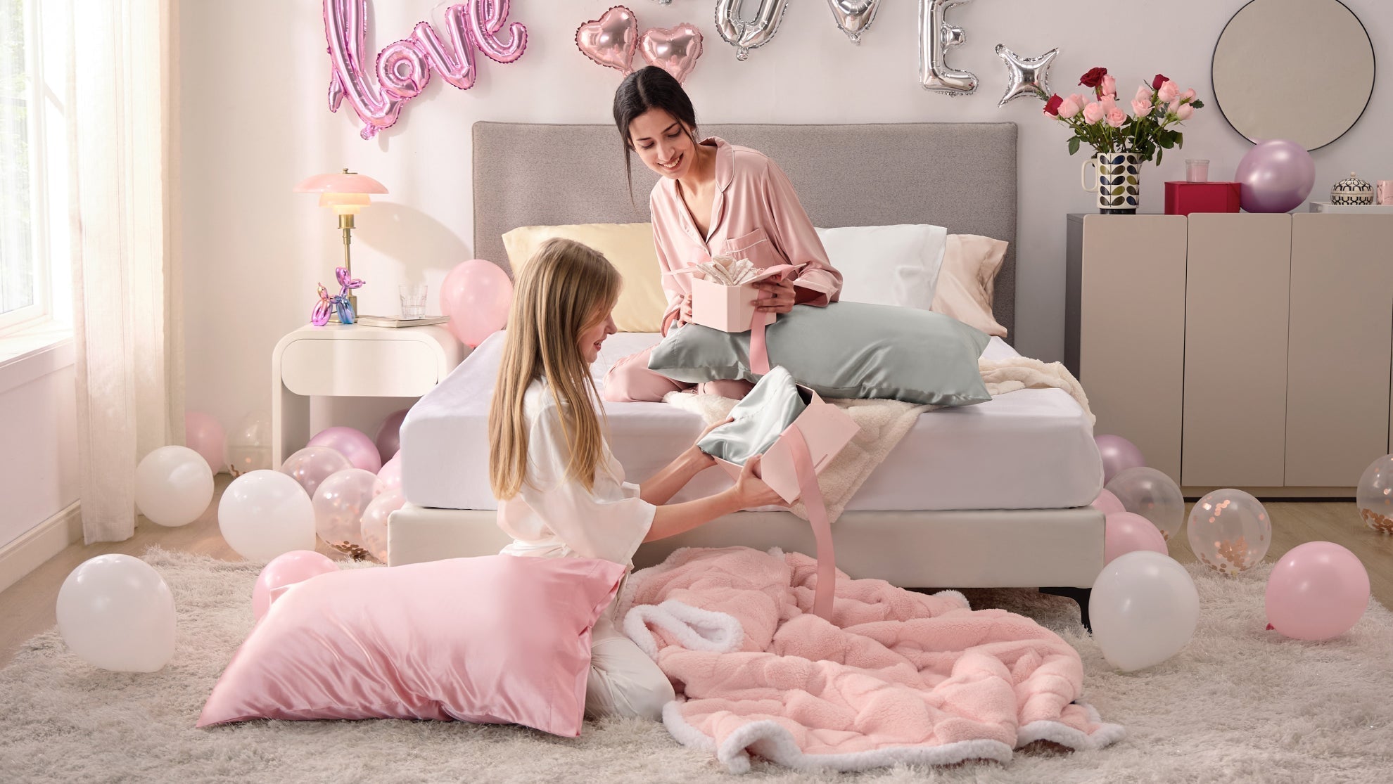 Two women in pajamas opening Valentine's gift boxes with luxury bedding on a bed decorated with pink balloons.