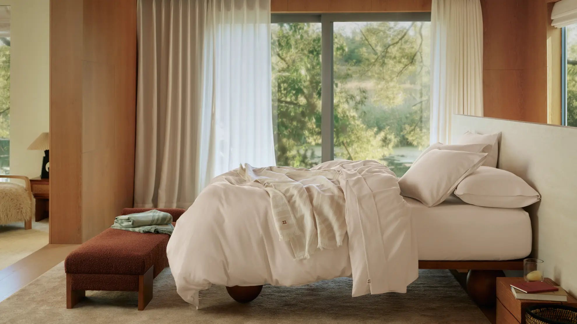 A sunlit, wood-paneled bedroom transition with airy cream bedding, a bed-end bench, and large windows overlooking lush greenery.