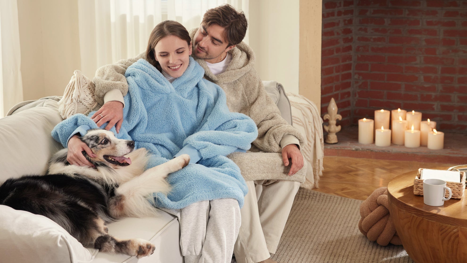 A happy couple wearing thick, oversized sherpa hoodie blankets relaxes on a couch with their dog, enjoying a cozy, warm night by candlelight.