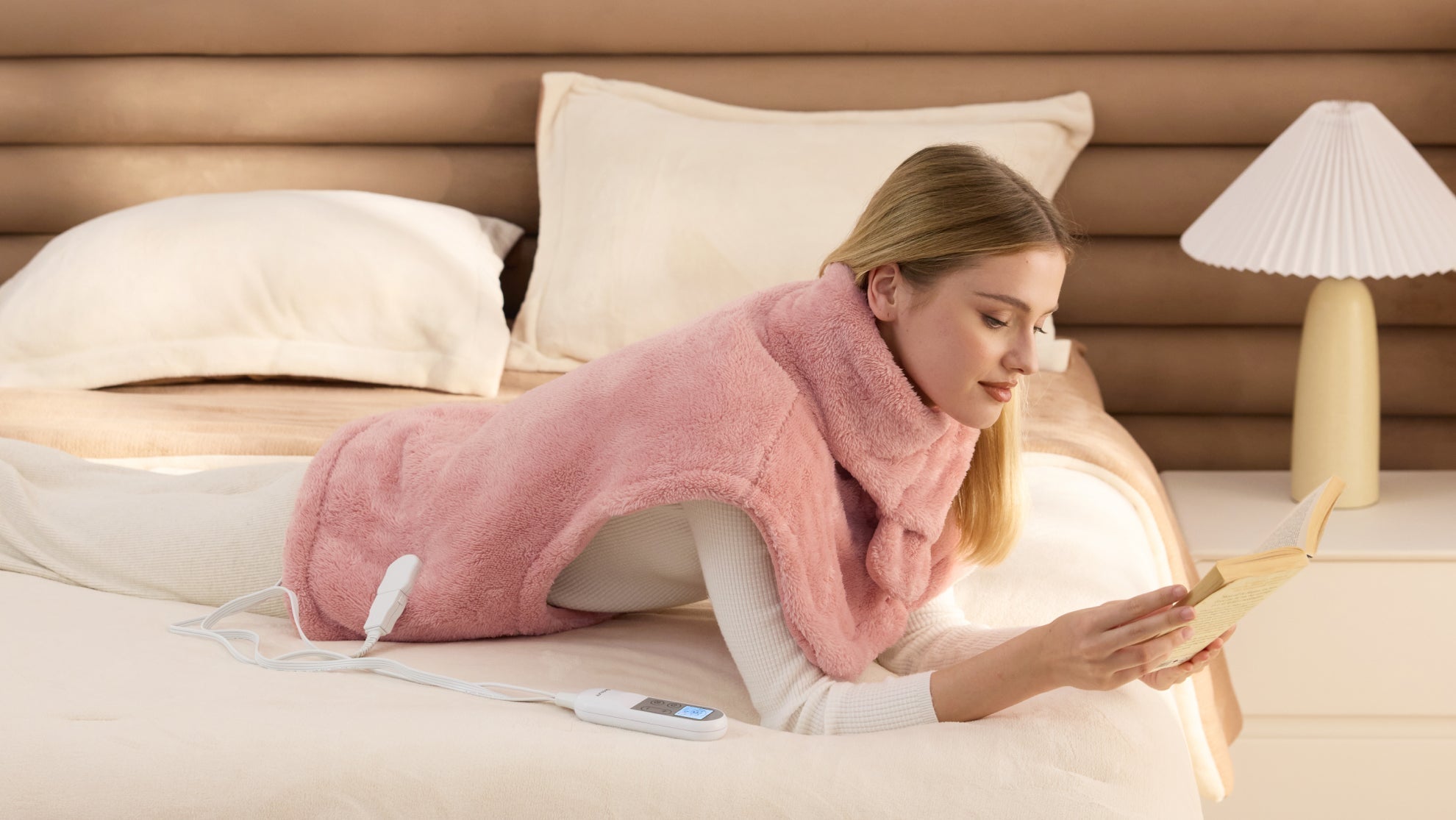 A woman using a pink heated pad on her shoulders while reading to relieve muscle tension and neck pain.