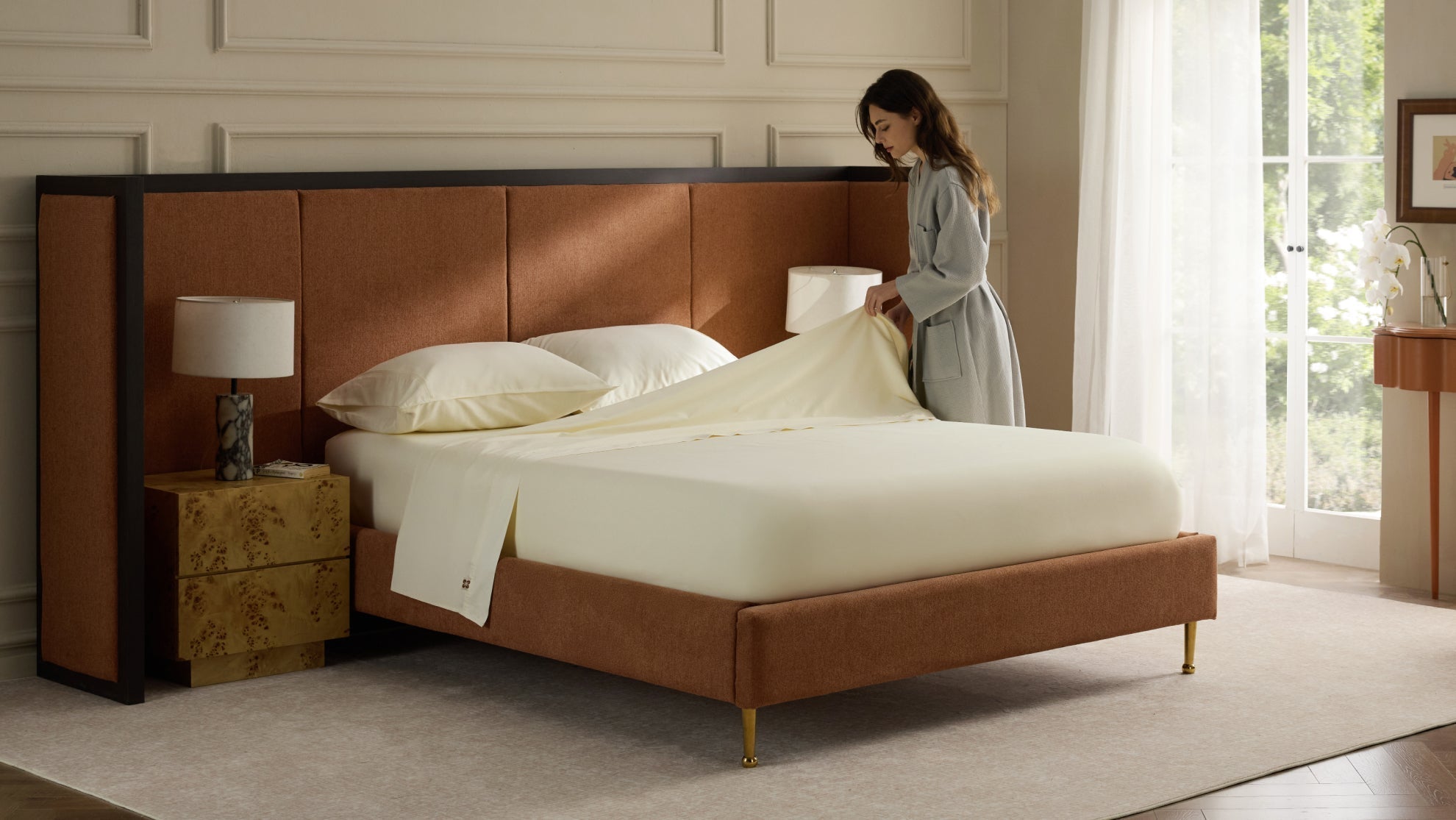 A woman in a grey robe makes a bed with off-white sheets in a modern, stylish bedroom.