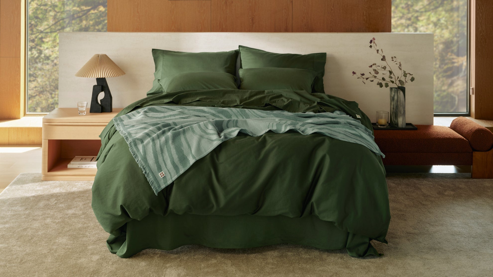 A luxurious bed is made with dark olive green bamboo sheets and a patterned throw in a modern, wood-paneled bedroom.
