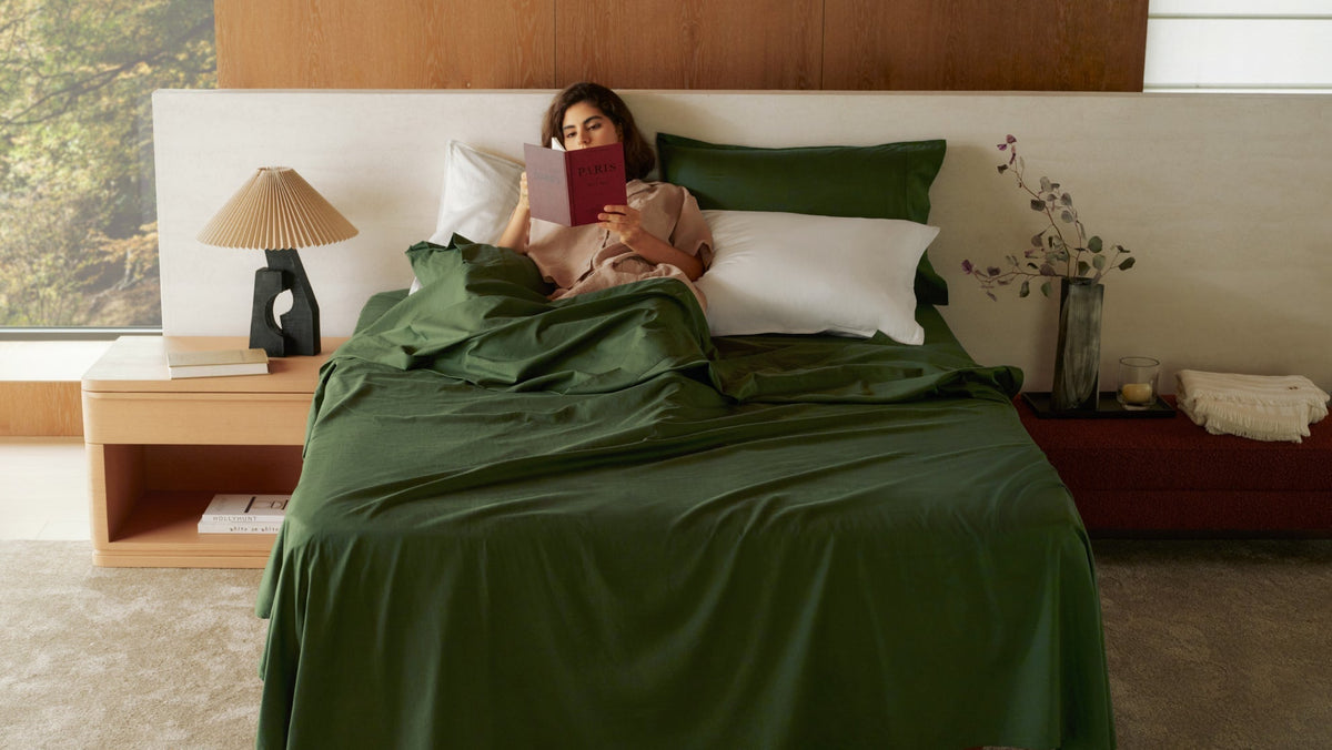 A woman reading comfortably in a bed with dark green sheets that stay in place during restful sleep.