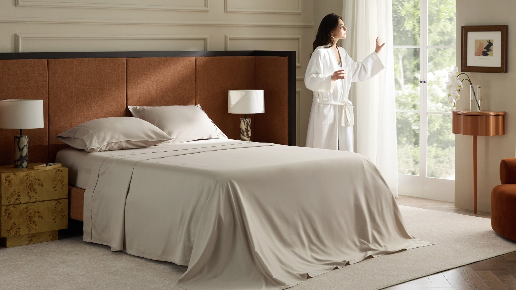 A woman in a white robe stands by a sunlit window in a bright bedroom featuring a large brown headboard, beige bedding, and elegant furniture.