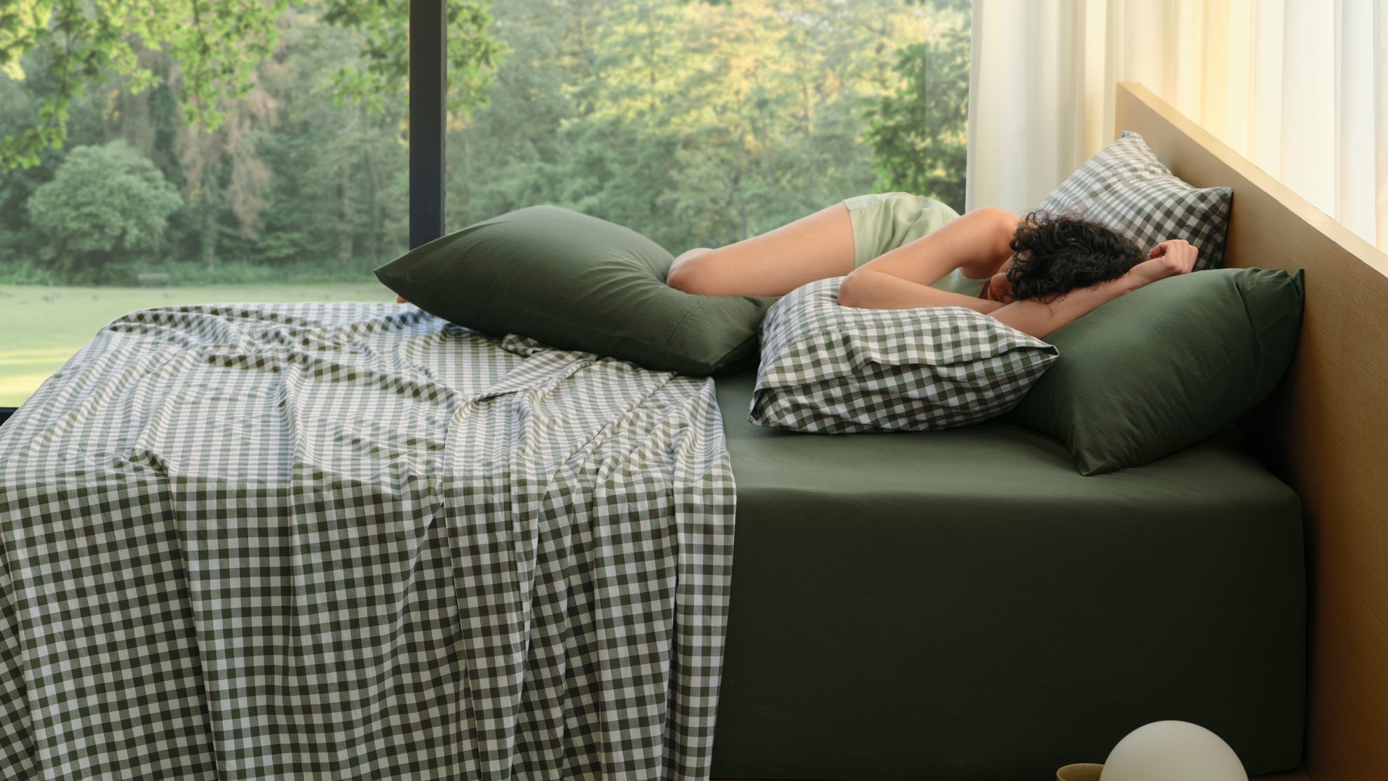 A woman rests on 100% Cotton Sheets featuring a green gingham pattern near a bright window.