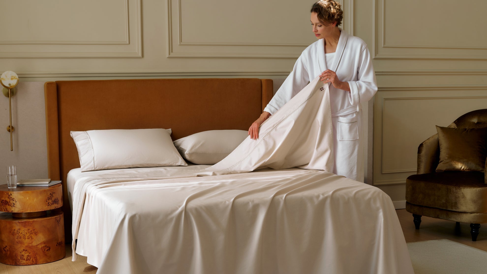 Woman adjusting smooth neutral linens on a minimalist bed in an uncluttered bedroom.