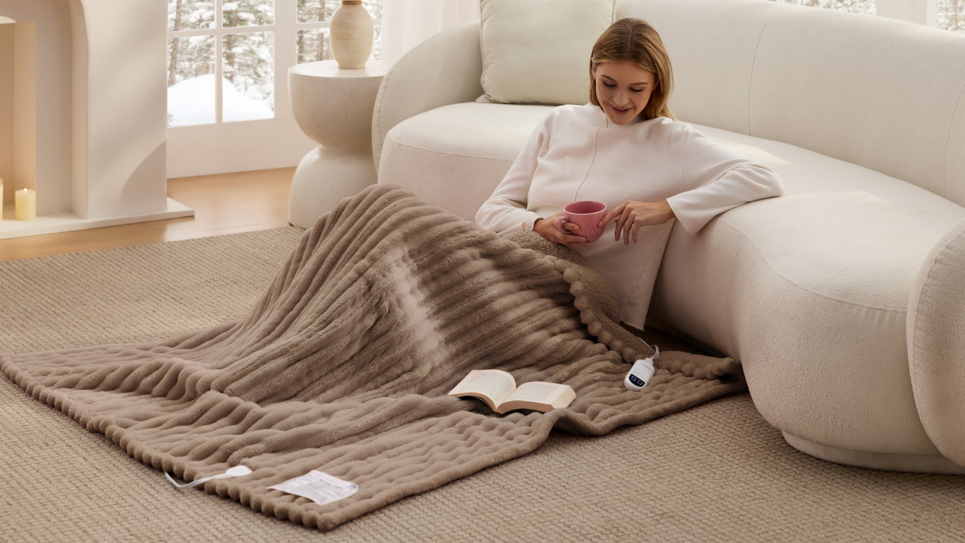 Smiling woman with a pink mug sits on a white sofa beside a draped blanket.