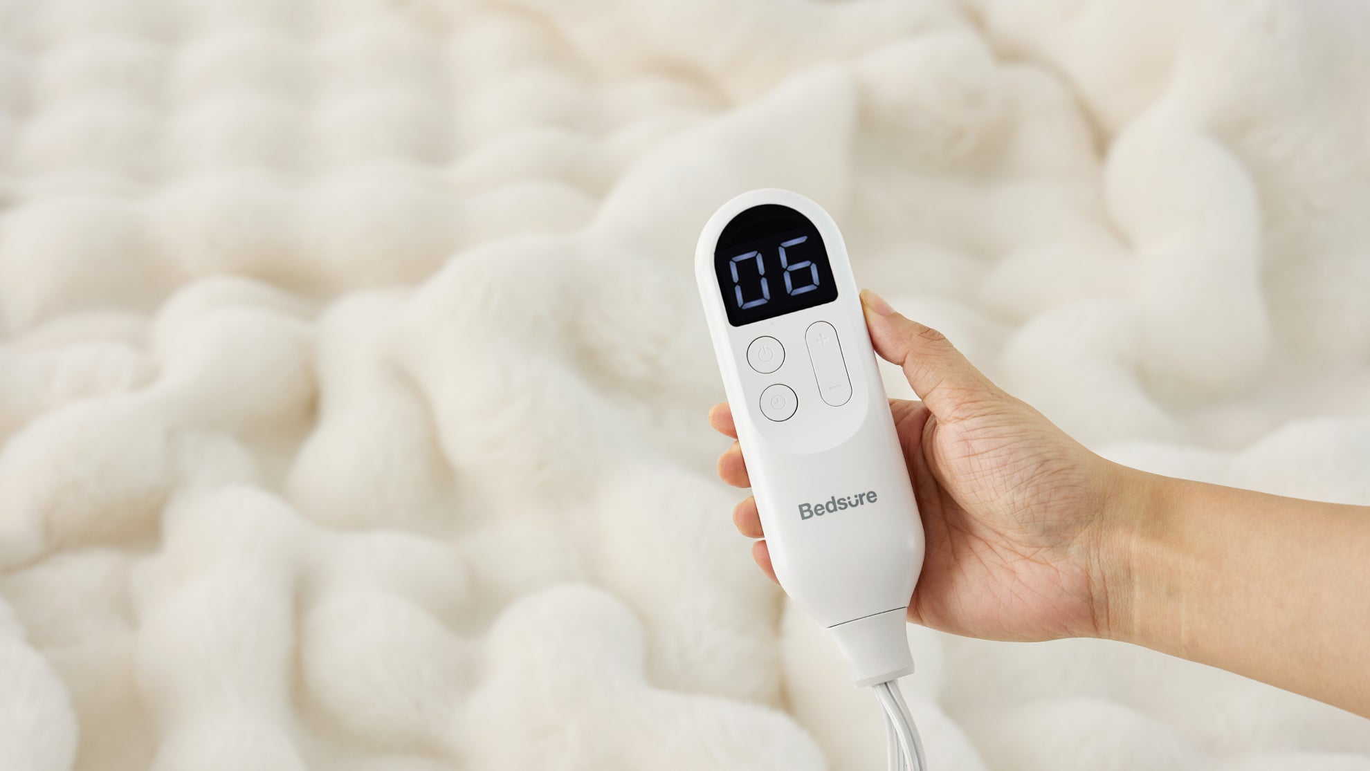 A hand holds a Bedsure controller above a fluffy white electric blanket, with the illuminated display showing a heat setting of 06.