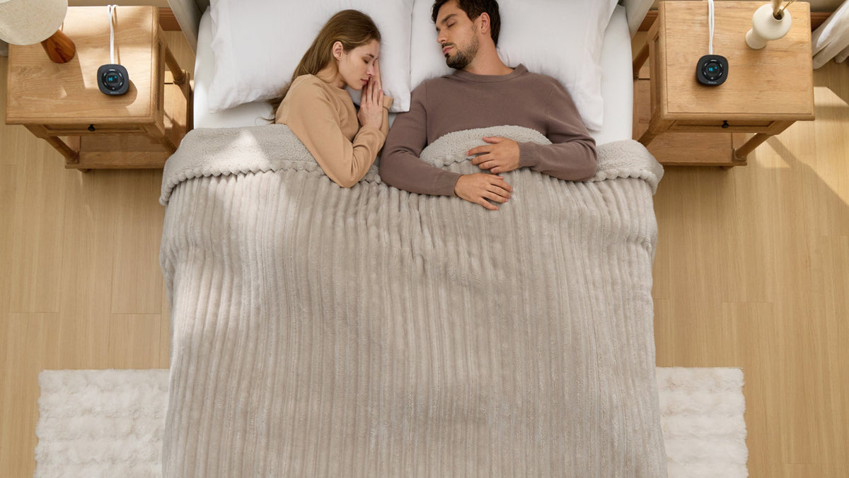 Overhead view of a couple lying side by side under a gray ribbed blanket, with separate controllers resting on their bedside tables.
