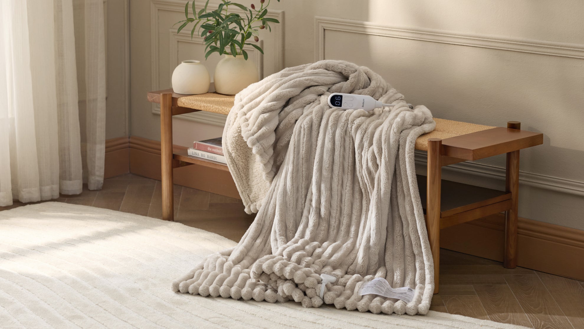 A gray ribbed electric blanket drapes over a wooden bench, with its white controller resting on top in a sunlit room.