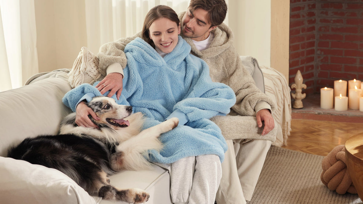 A couple in cozy fleece robes sits together on a sofa, smiling while petting a dog beside a candlelit fireplace.