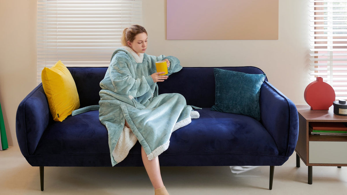 A woman wearing a light blue plush hooded robe sits on a navy sofa, holding a yellow mug beside colorful cushions and a wooden table.