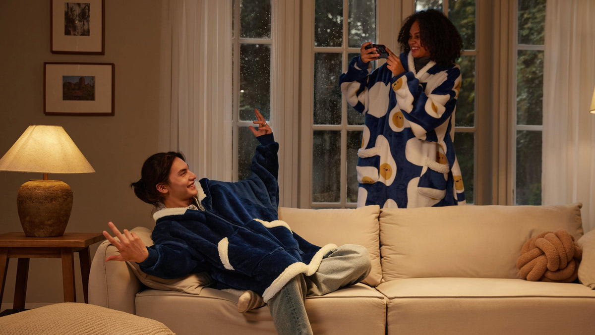 Two people on a cream sofa at night, one in a navy blanket hoodie with Sherpa trim and one in a blue printed hooded blanket holding a camera