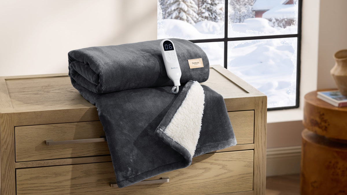 A folded grey heated blanket with a sherpa lining and its controller resting on a wooden dresser beside a snowy window
