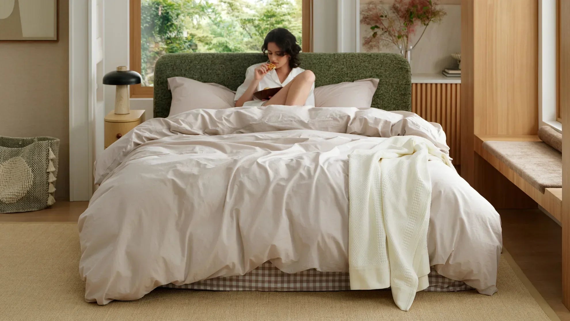 Woman relaxing in a bed layered with soft neutral bedding and a white throw