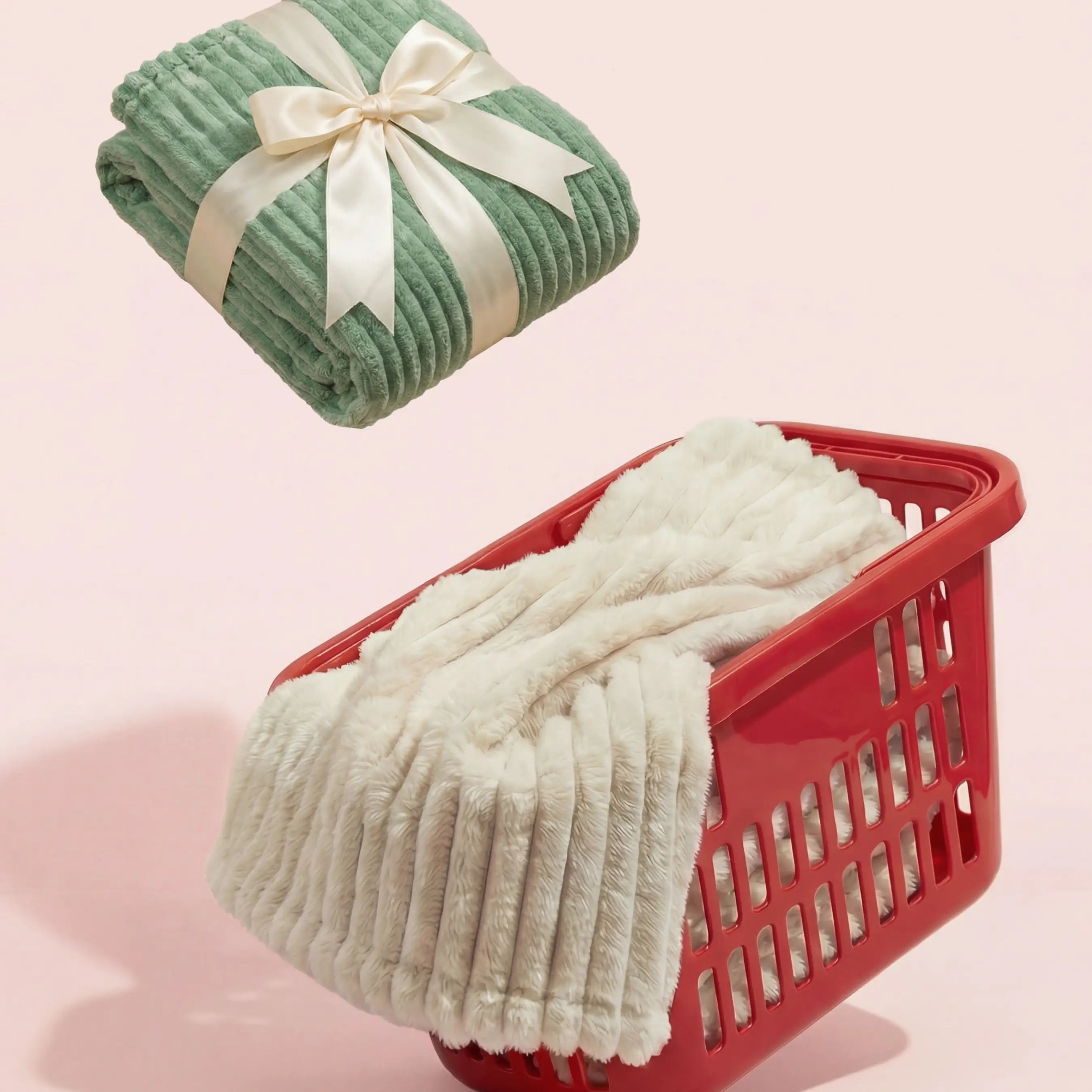 A sage green vertically ribbed plush blanket, wrapped and tied with a white satin ribbon bow, floats above a red plastic shopping basket containing an off-white vertically ribbed plush blanket, against a solid light pink background.