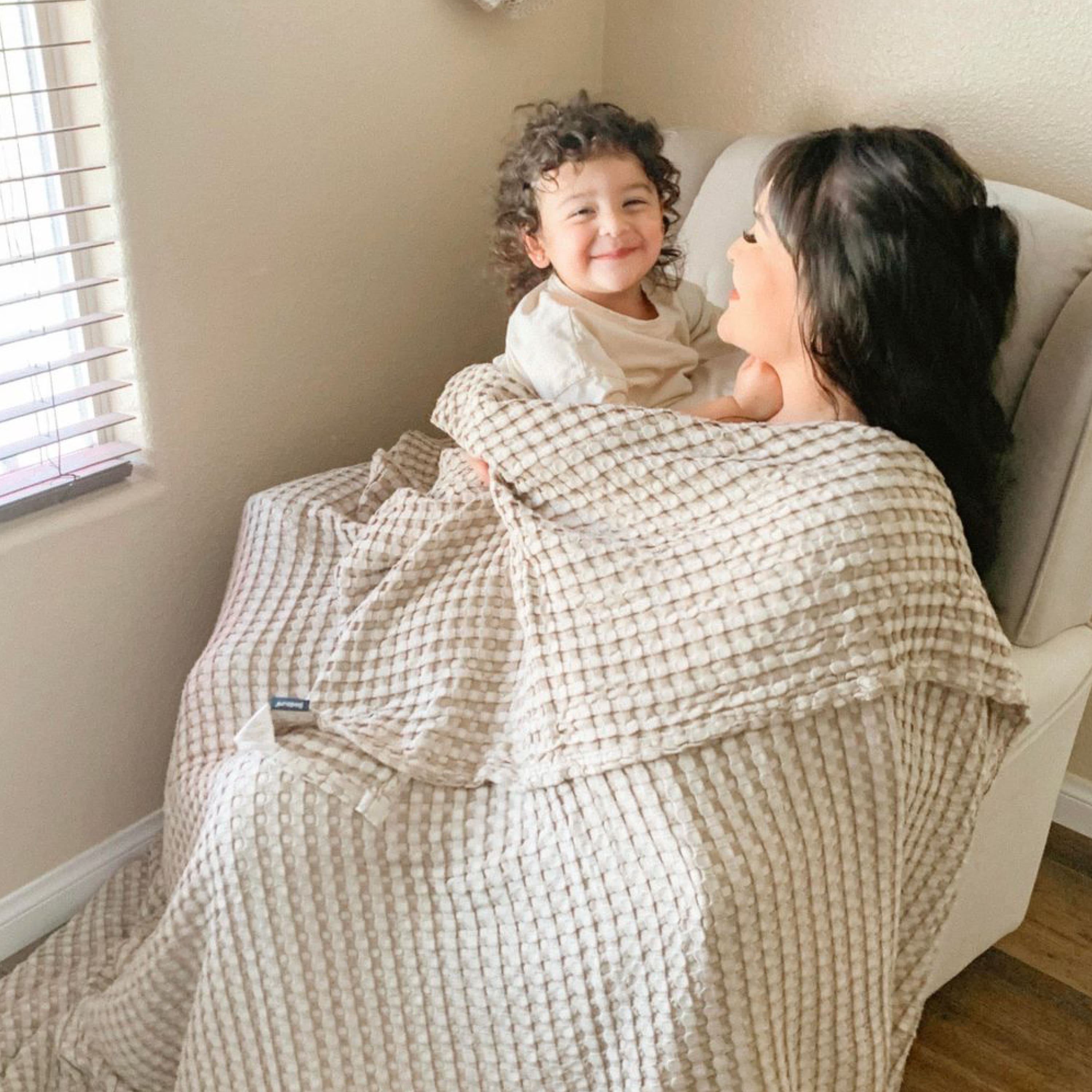 Woman and child wrapped in a beige textured blanket in a cozy room. 