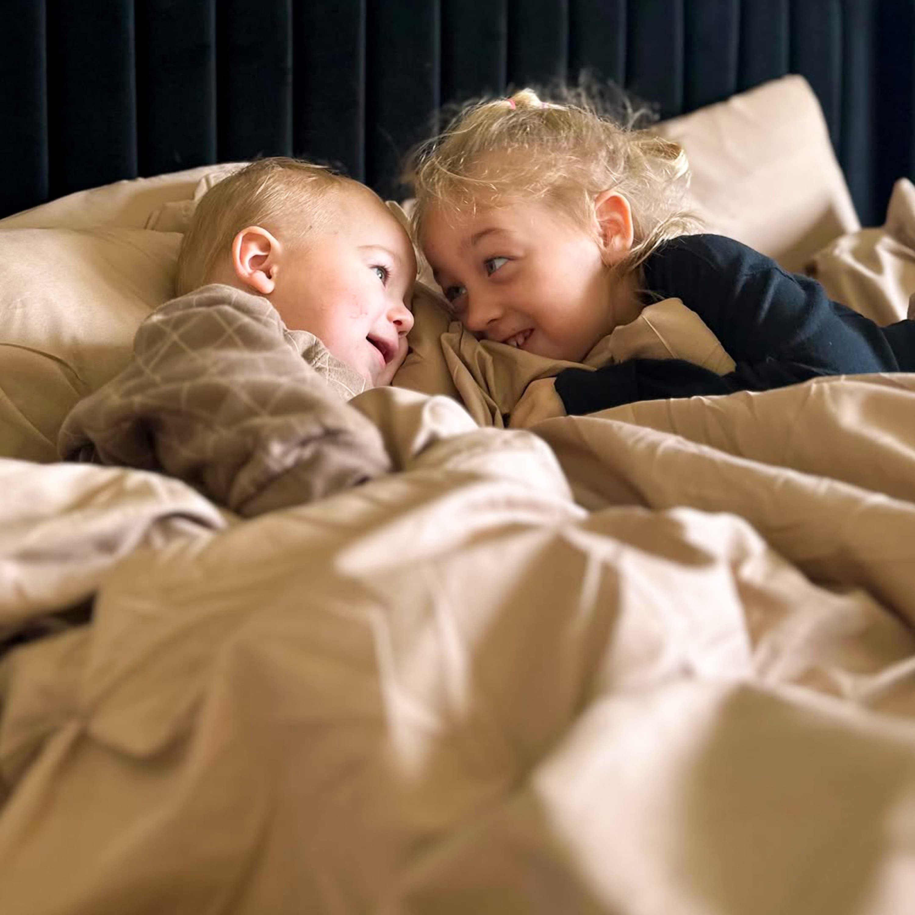 Two children lying in bed under a blanket, looking at each other. 